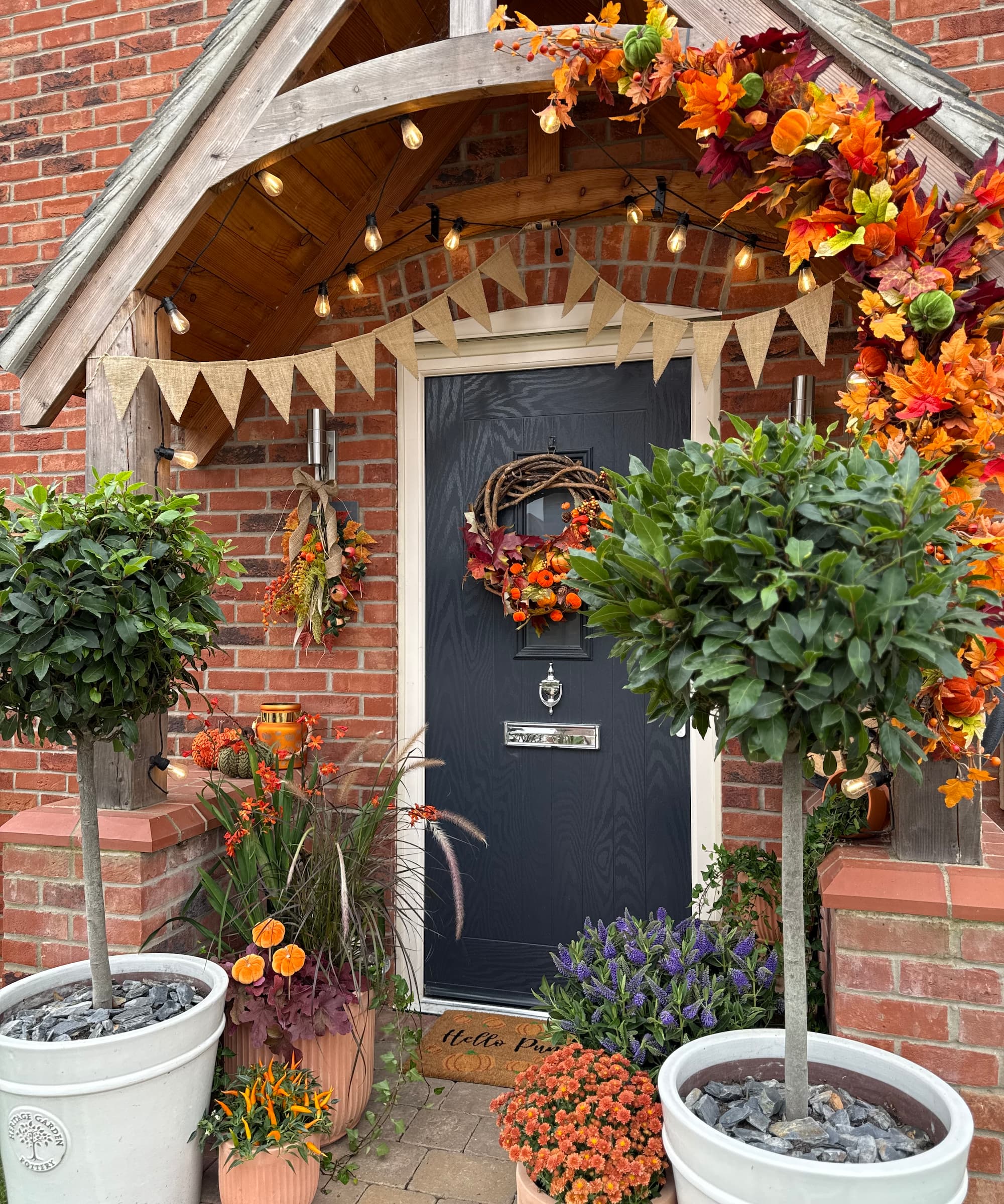A front door with two bay leef trees and a black door with lighting and a porch above