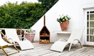 Corten steel fire pit with lit logs, wicker loungers, terracotta pots and white flowers on wooden deck.