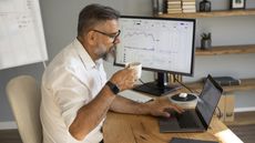 An older man looks at his laptop while sitting at his desk at home, a trading website on the monitor beside him.