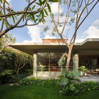 Daytime exterior side image of Ventana House in Mexico among greenery, flat white roof, red brick chimney, green lawn, shrubs, plants and trees, windows, stone patio area, tables and chairs, blue sky with light white clouds