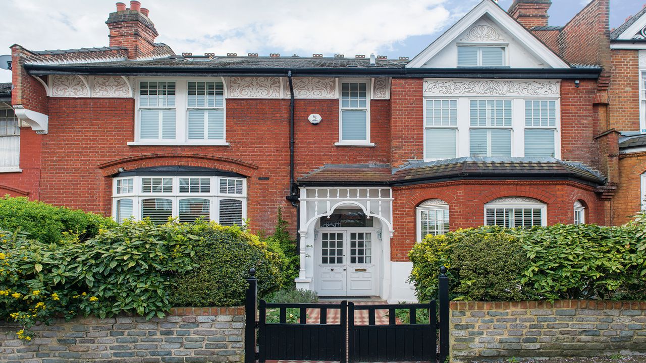 red brick house exterior with black front gate, white windows and doors and green hedges above a stone wall