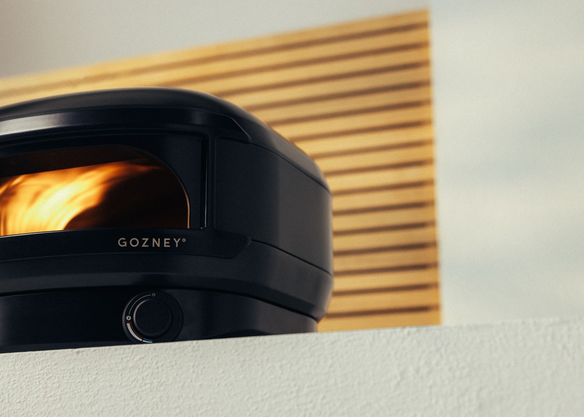 Gozney's Arc Lite pizza oven in off-black against a wood-slat divider screen and otherwise cream background