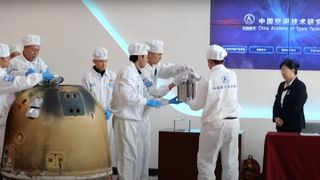 seven people in white lab anti-contamination suits remove a small silver box from a charred bronze-colored space caspule inside a white-walled room