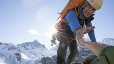A mountain-climbing older man reaches a hand out to an unseen climber to help him up.