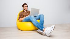 A cheerful-looking man sitting on a beanbag chair and working on a laptop.