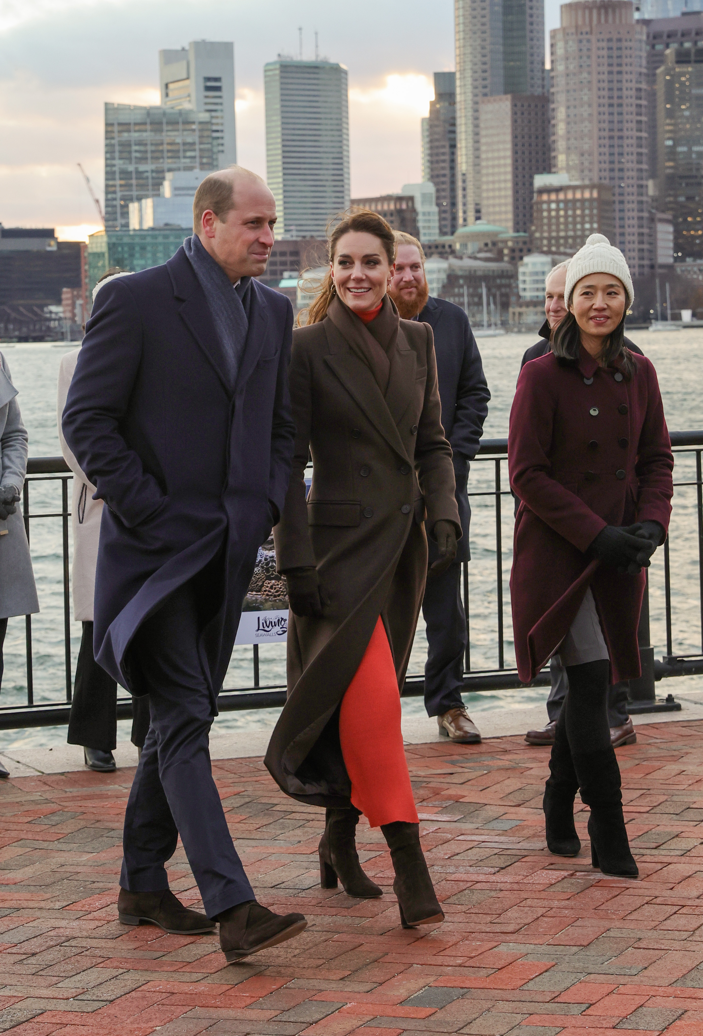 Kate Middleton walking in an orange skirt and brown coat next to Prince William and a group of people along the Boston skyline and shore