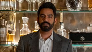 Sunil wearing a blazer and holding a glass of liquor while in front of a large bar full of alcohol bottles in Death and Other Details