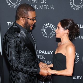 Tyler Perry wears a black suit featuring a shiny pattern while holding hands with Meghan Markle, who wears a black strapless dress