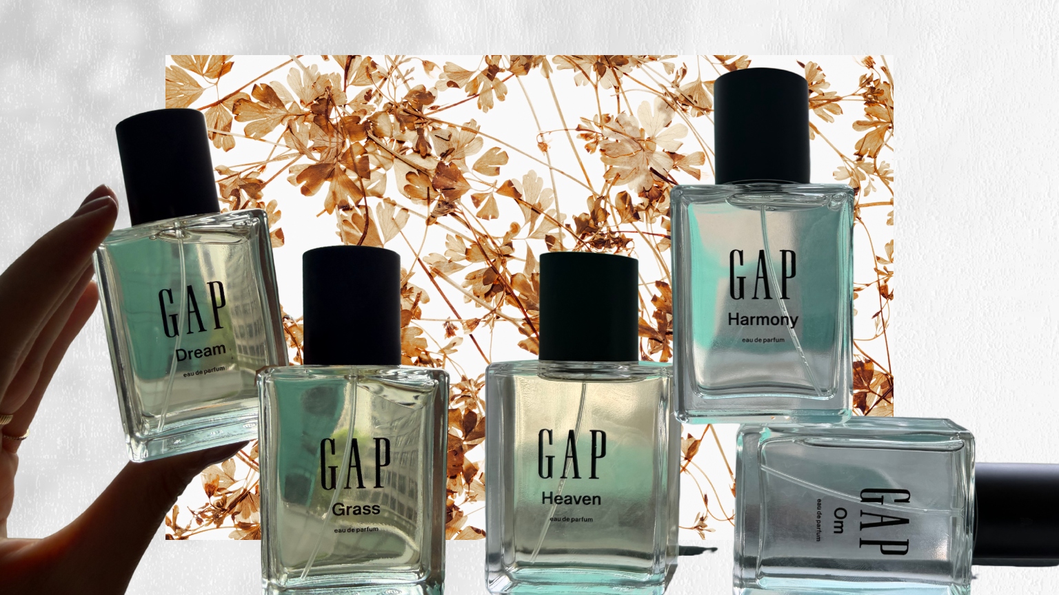 a white shadow background with brown and gold leaves under a hand holding a glass bottle of Gap perfume next to four other glass bottles of Gap perfume