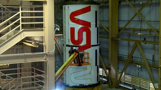 a rocket standing up with a nasa logo half-painted. people on a crane wield spray paint. the rocket is in a warehouse. the door is visible at right and a large staircase visible at left