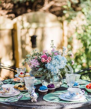 An outdoor dining area with a round black metal table with green placemats, colorful vintage teacups, glasses, pastries and cupcakes, and a glass vase in the middle with blue, purple, and pink flowers