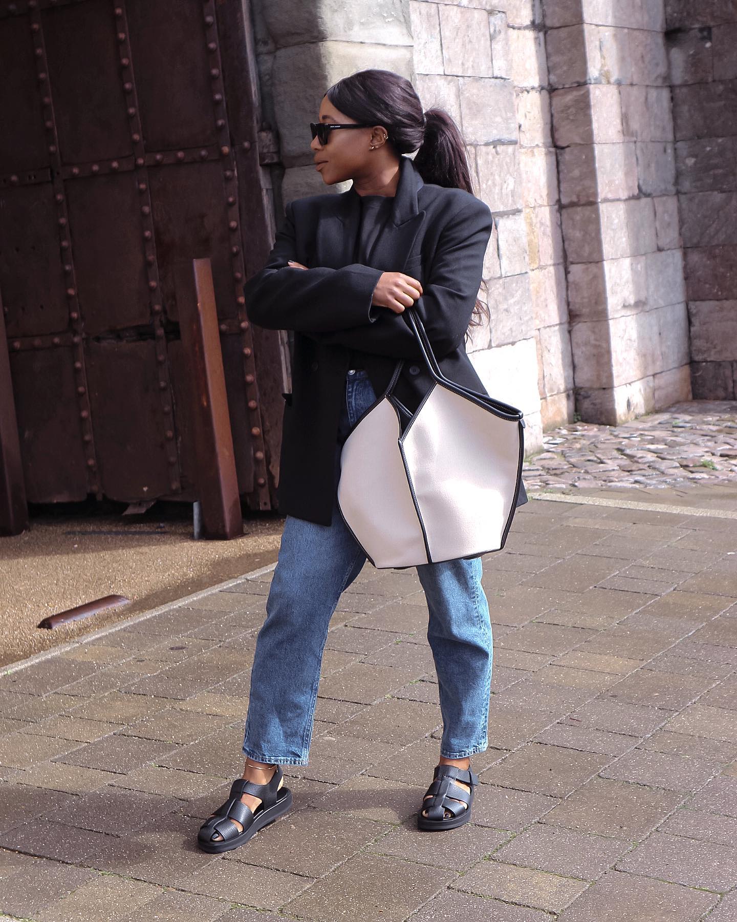 Woman wearing a black blazer, blue jeans, and black sandals and carrying a white tote bag..