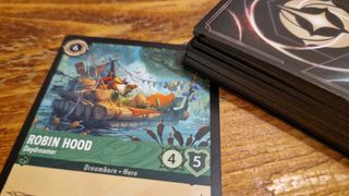 Robin Hood relaxes on a raft in artwork from a Disney Lorcana Into the Inklands card, sat on a wooden table beside a deck