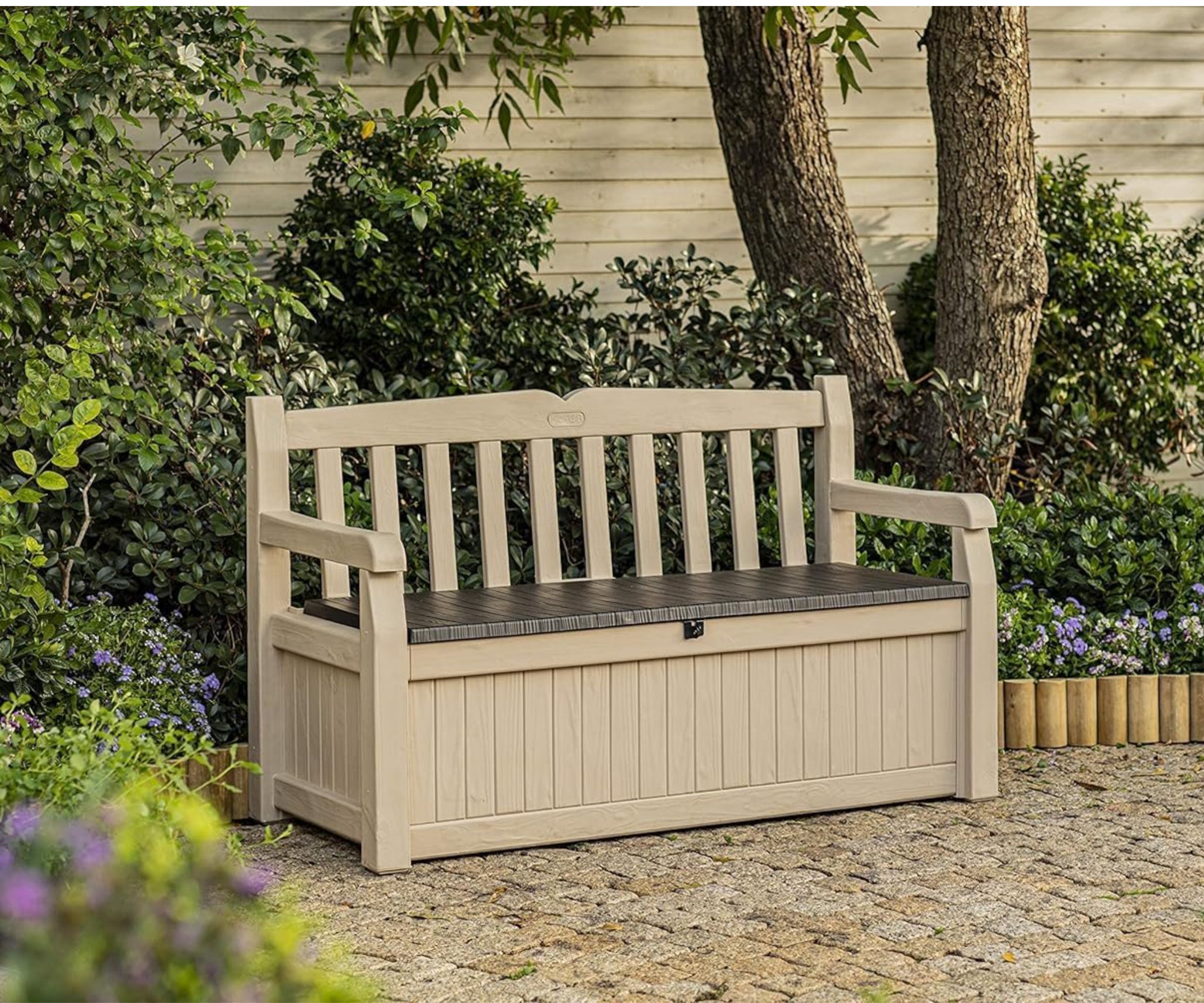 Beige outdoor storage bench with a dark brown seat, surrounded by lush greenery and flowers.