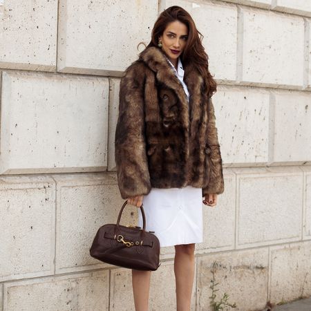 A woman wears a brown short fur coat, a white mini dress, and a brown top handle bag.