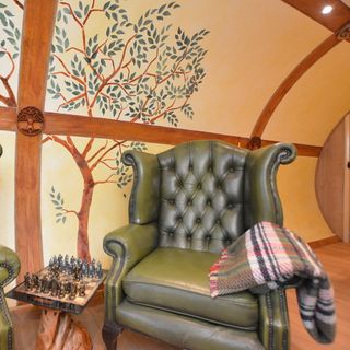 a green leather Chesterfield armchair with a tartan blanket resting over the arm, next to a wooden side table which has a chessboard on top, in front of the curved yellow wall with wooden panel beams and tree design