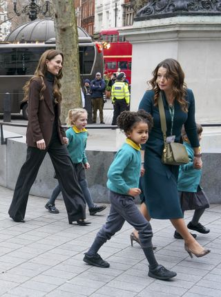 Kate Middleton holding hands with a little girl in a teal jumper walking behind a teacher and two other students on a London street with a bus behind them