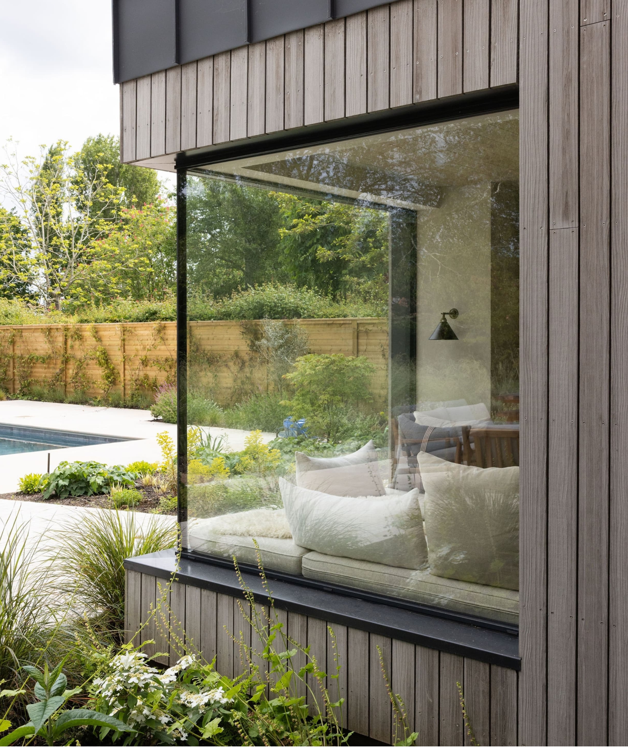 Frameless glass corner window overlooking a garden and pool, framing a cushioned window seat inside.