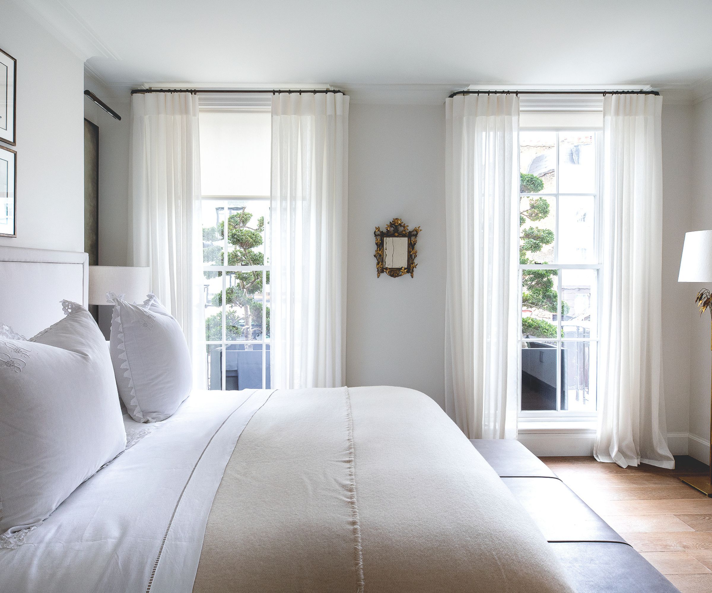 All white bedroom with sheer drapes, neutral bedding, wooden floors and gold mirror