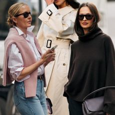 A woman in straight-leg jeans, a button-down, and a pink sweater over her shoulder. Woman in a trench coat at London Fashion Week S/S 26. A woman in a brown oversized turtleneck and black trousers. 