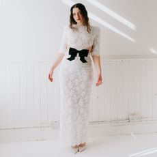 Lucy Halliday photographed by Phill Taylor. She is in a white room wearing a white lace dress with center cutout and black bow. 