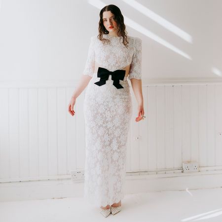 Lucy Halliday photographed by Phill Taylor. She is in a white room wearing a white lace dress with center cutout and black bow. 