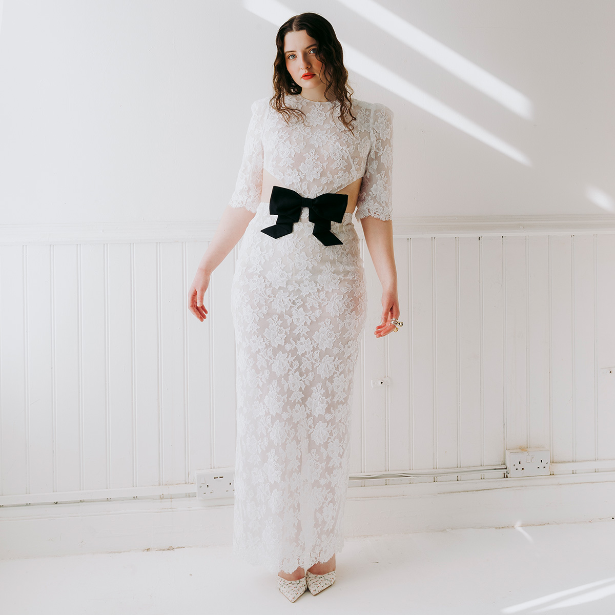 Lucy Halliday photographed by Phill Taylor. She is in a white room wearing a white lace dress with center cutout and black bow. 
