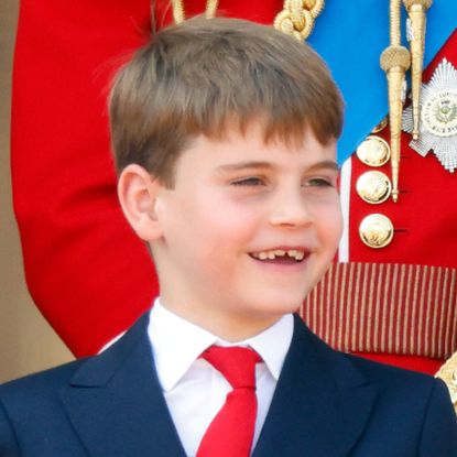 Prince Louis smiles, showing off his growing teeth, while wearing a navy suit with a white shirt and red tie