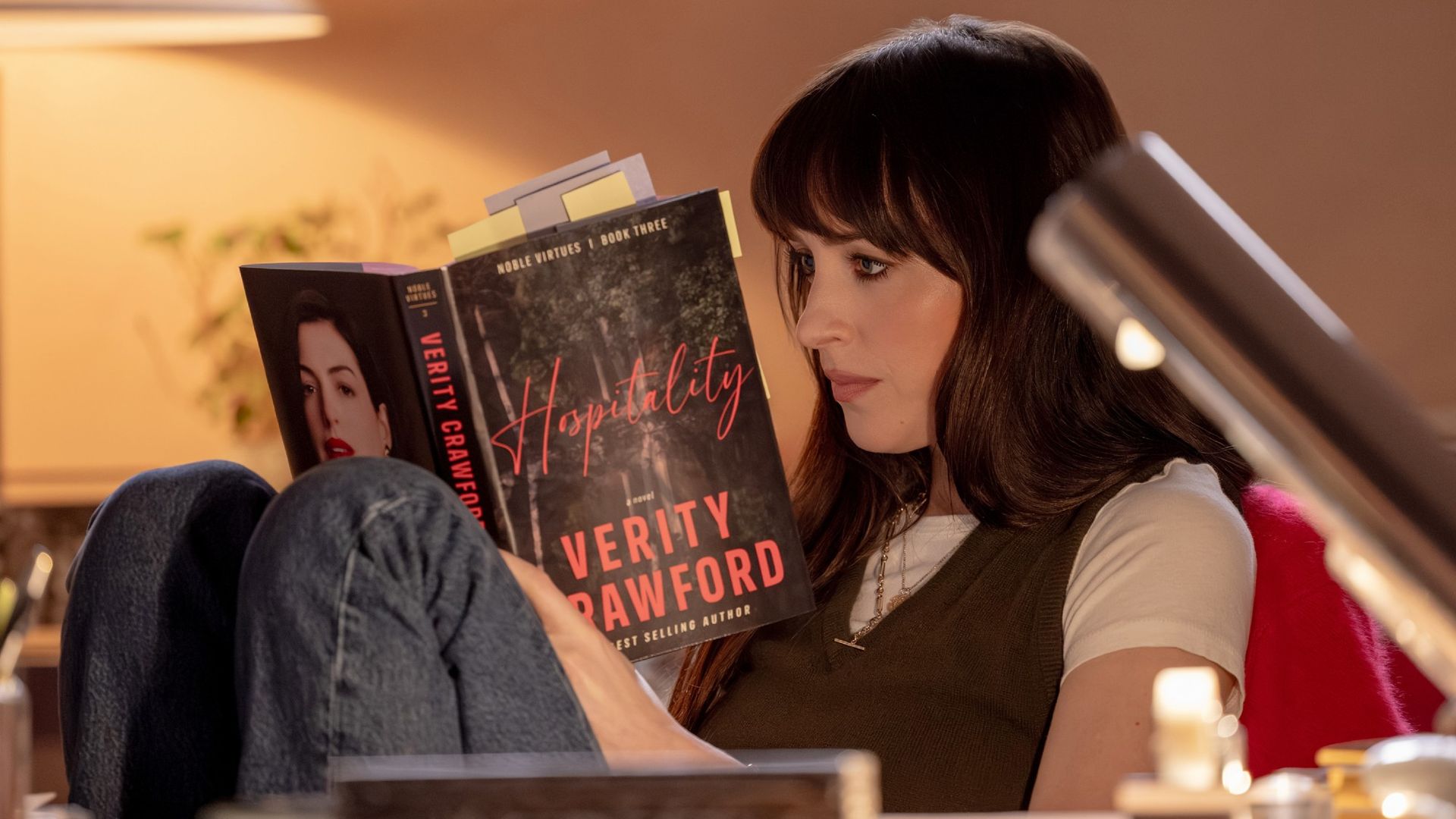 Dakota Johnson as Lowen reading a book with Anne Hathaway's Verity on the back cover.