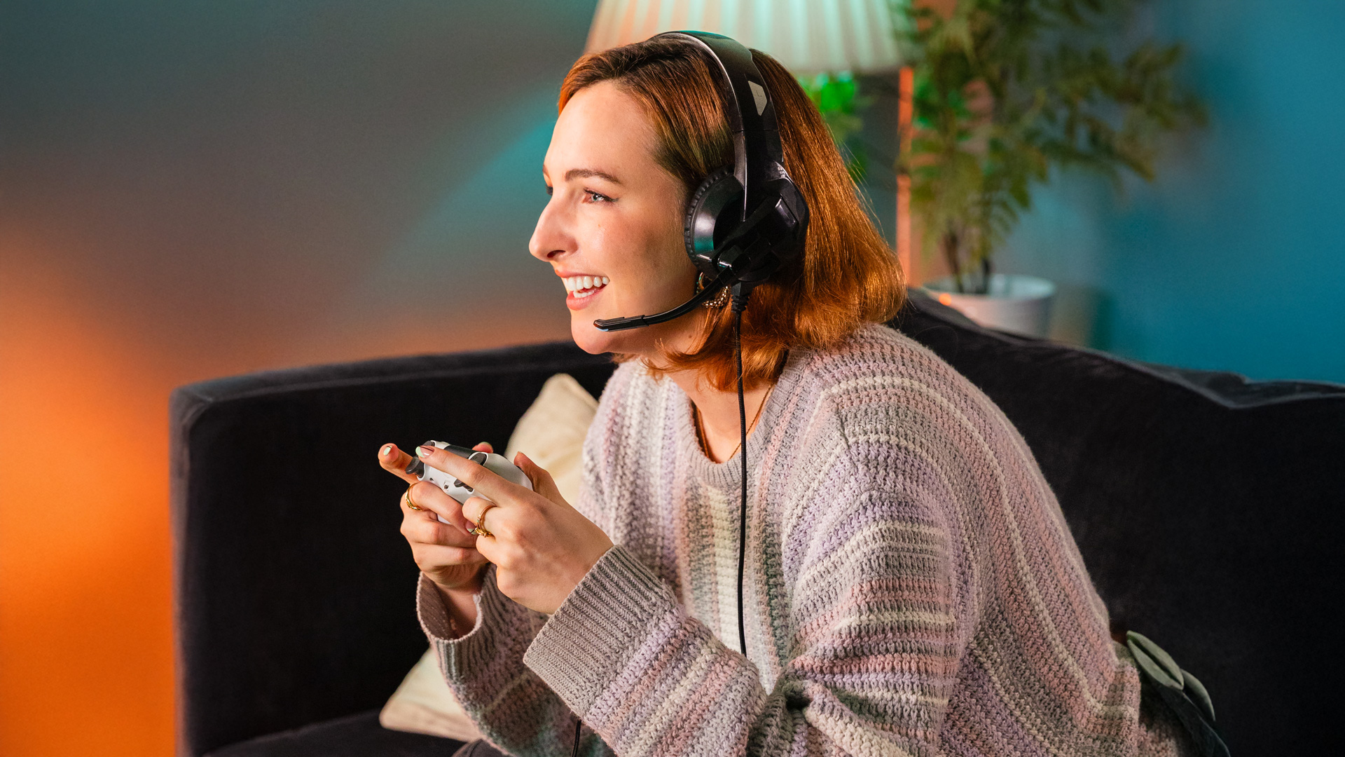 A young woman sits on a sofa wearing a gaming headset and holding a PS5 controller