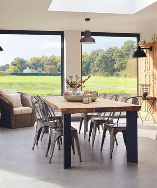 Dining room area with full length windows out onto the garden, white walls and grey polished tiled floor, skylight and wooden table and metal chairs.