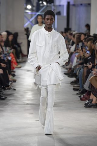 A female model wearing a white long sleeved button up shirt and white bell bottom pants walking down a runway.