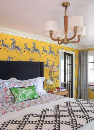 yellow bedroom with yellow animal design wallpaper, black bed, floral bedlinen, black and cream blanket, grey drapes, green pillow cushion