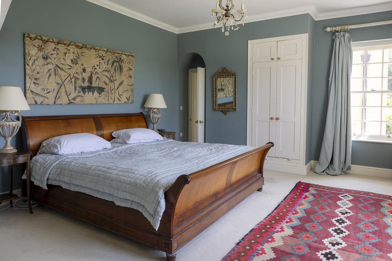 A bedroom complete with brown bed frame and light blue bedding and a red rug with a diamond pattern on it inside Mount Farm