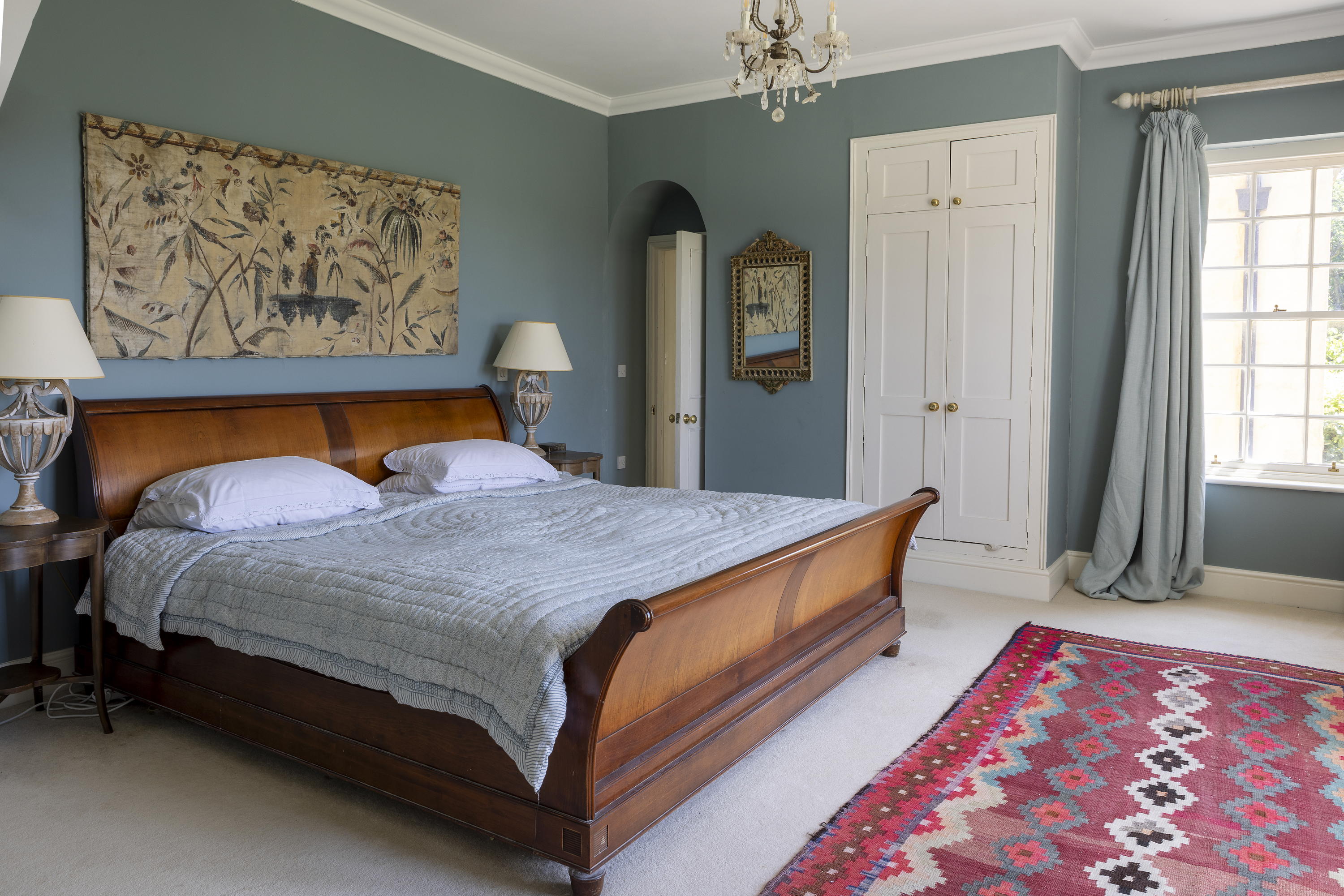 A bedroom complete with brown bed frame and light blue bedding and a red rug with a diamond pattern on it inside Mount Farm