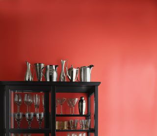 A black glass fronted cabinet against a red wall with glassware inside and a metal jugs on top