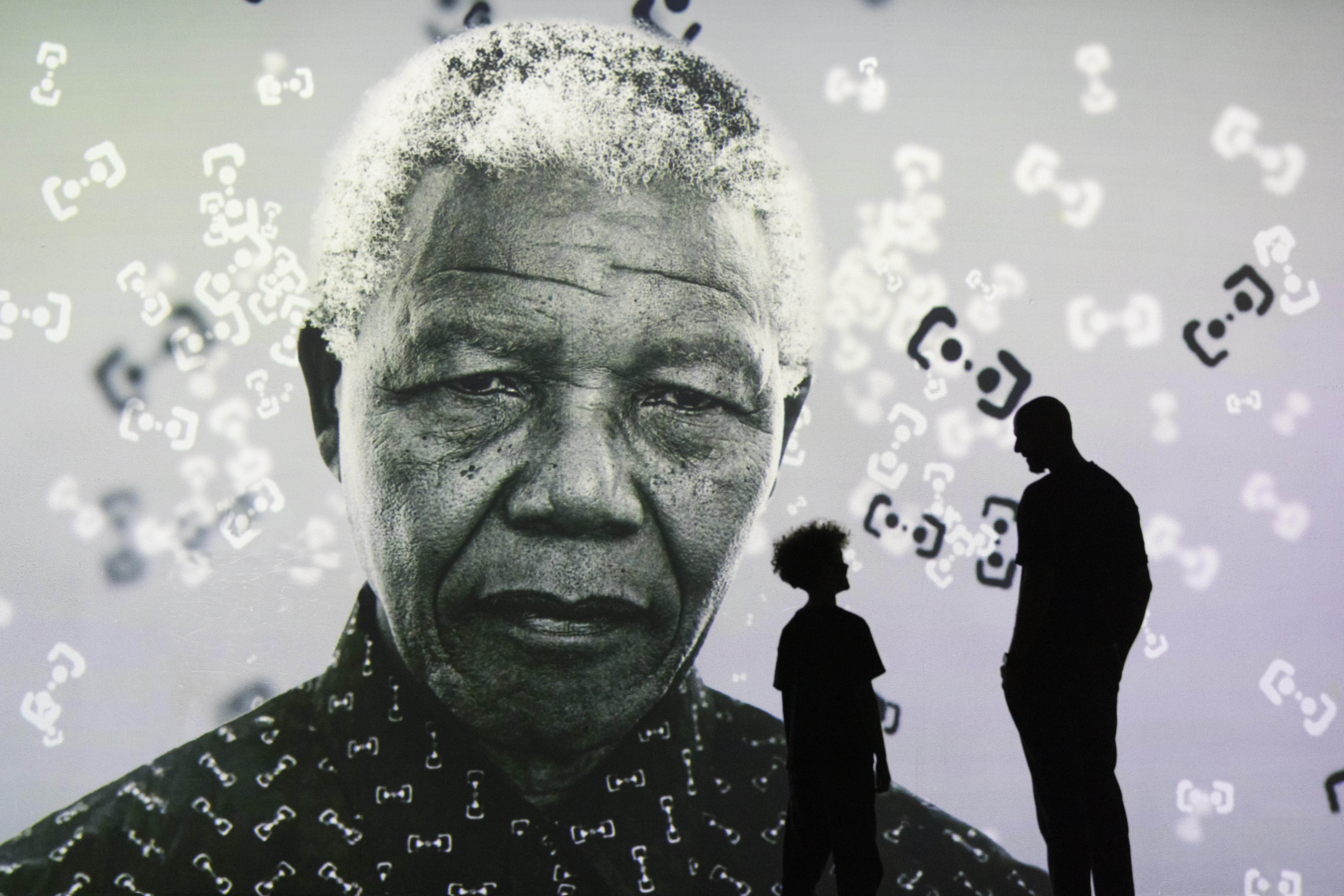 The silhouettes of an adult and a child stand before a vast black-and-white projection of Nelson Mandela's portrait at FRAMELESS London, with floating abstract symbols scattered across the image.