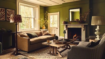 A deep green living room with two sofas, a wooden table in the center. A large stone fireplace with a lit fire inside. 
