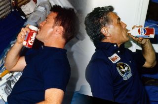 NASA astronauts Tony England (at left) and Karl Henize, STS-51F mission specialists, drink from Coca-Cola and Pepsi space cans as part of the Carbonated Beverage Dispenser Evaluation on space shuttle Challenger in 1985.
