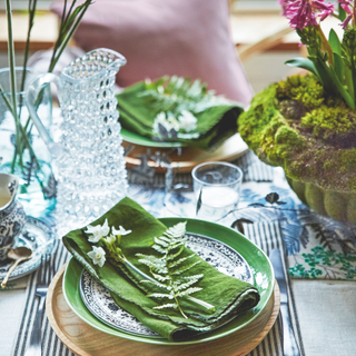 Place setting with fern frond and flowers on green napkin beside flower arrangements.