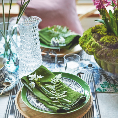 Place setting with fern frond and flowers on green napkin beside flower arrangements.