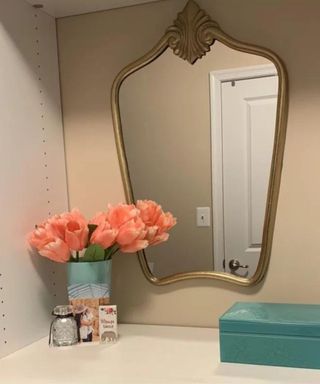 A white unit with a gold rimmed mirror and a vase of flowers