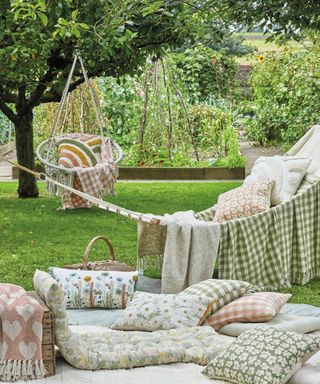 A garden area with a white hammock and hanging chair adorned with green, pink, and white floral pillows and throws, and a picnic area on the lawn with pillows, throws, and a basket
