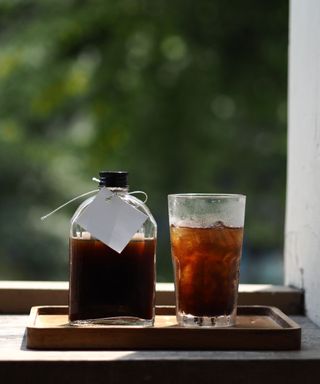 A glass cup of brown cold brew coffee with a glass bottle filled with cold brew to the left of it, stood on a wooden board on a wooden ledge, with green leaves beyond the window