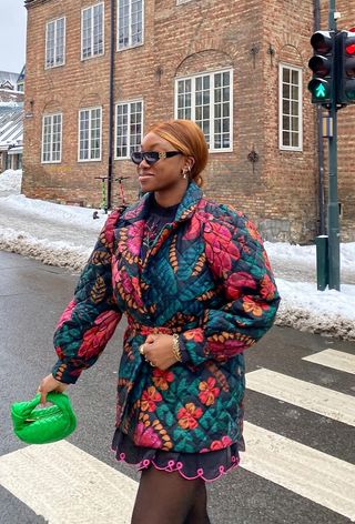 quilted patchwork jacket trend is shown on a woman walking across the street wearing a printed floral jacket over a dress with tights and a green bag