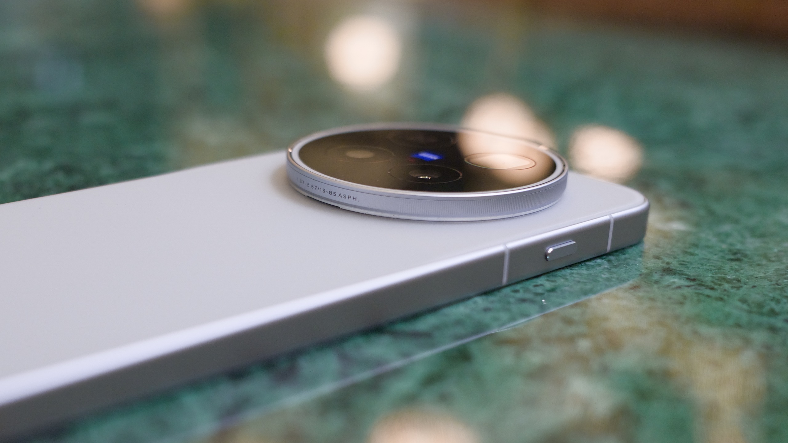 Low-angle close-up of the Vivo X500 Pro Max lying on a green marble table, emphasizing the raised circular Zeiss camera module and flat metal frame.