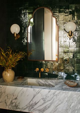 Marble farmhouse bathroom sink backed with reflective green tiles and large brass mirror