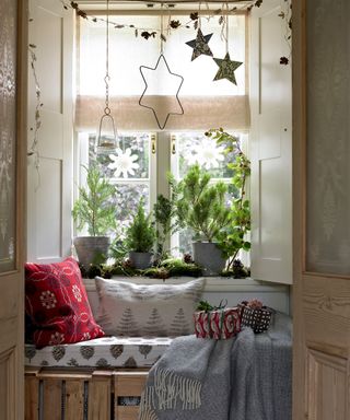 Window seat covered with christmas decorated pillowcases and gifts in front of planted trees and a white window