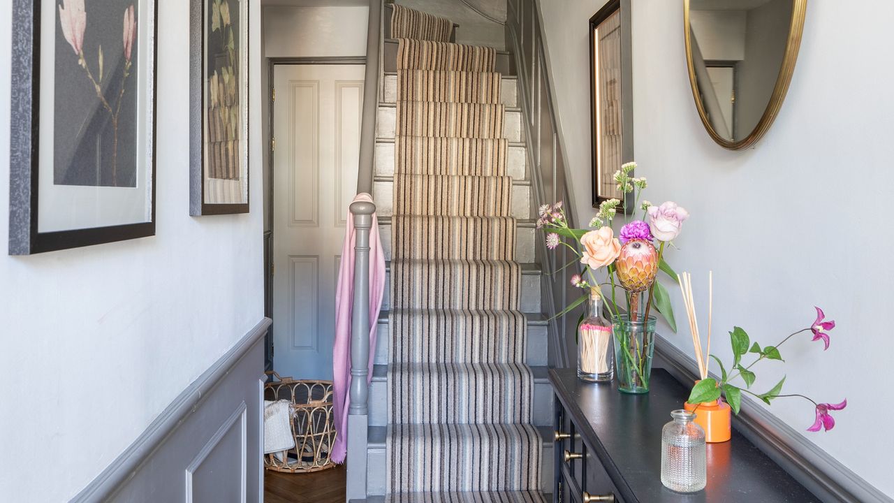 A narrow hallway half painted in dark grey and half in white with a staircase at the end and a storage console table decorated with flowers to the side