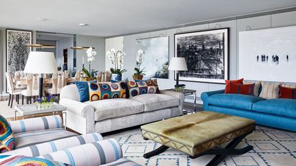  10 reasons why your friends hate your house. Living room with tan trestle coffee table flanked by white, blue and striped sofas with accent red and retro patterned cushions, and open plan dining area beyond